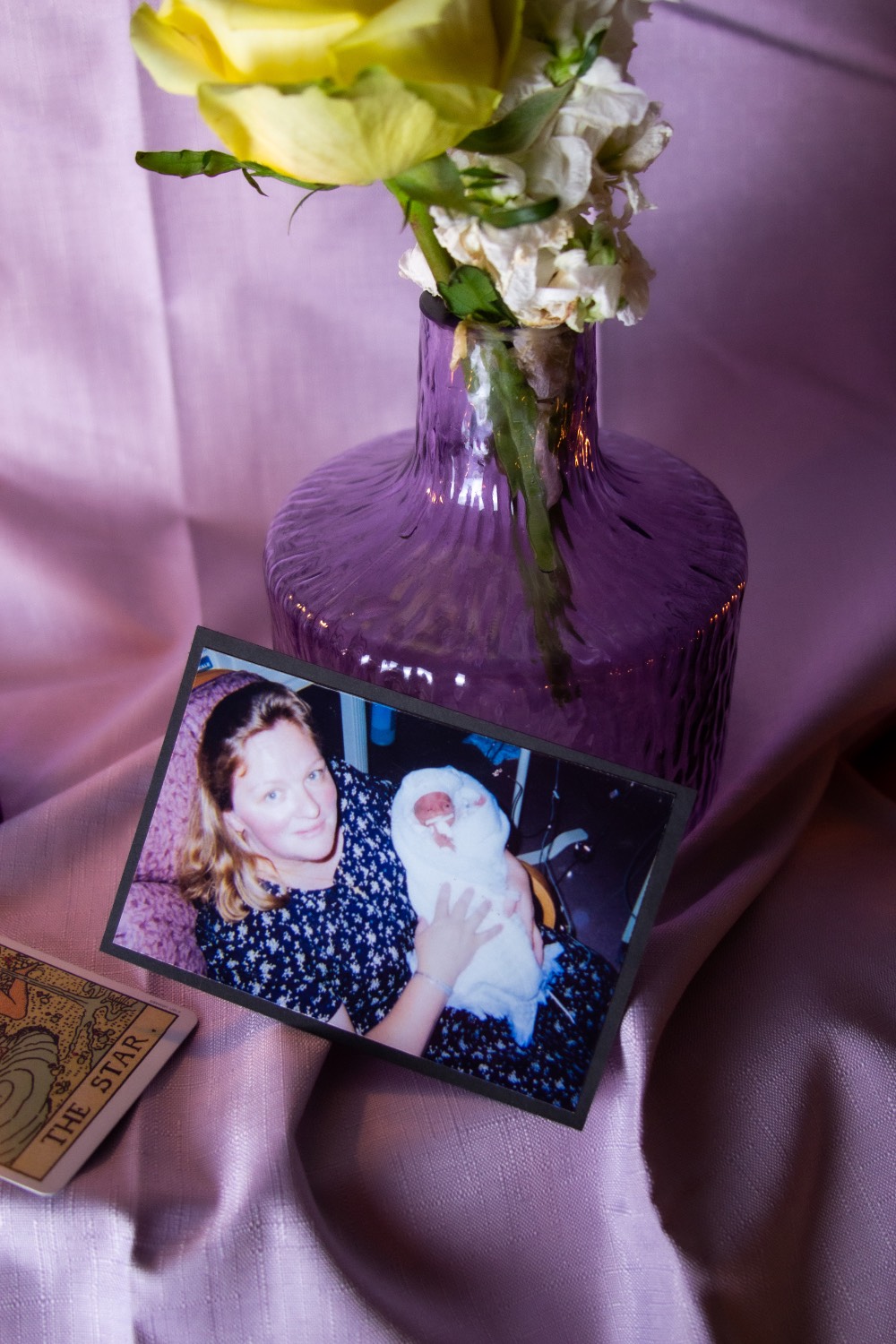Photo of a photograph of a mother holding a baby. The photo sits on a purple cloth against a purple vase holding a yellow flower.